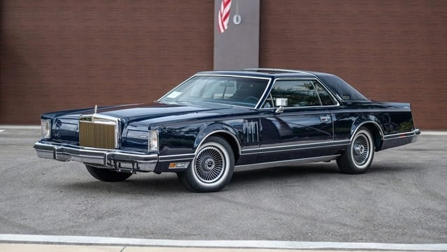 Sudut kiri depan mobil coupe biru tua yang diparkir di aspal.