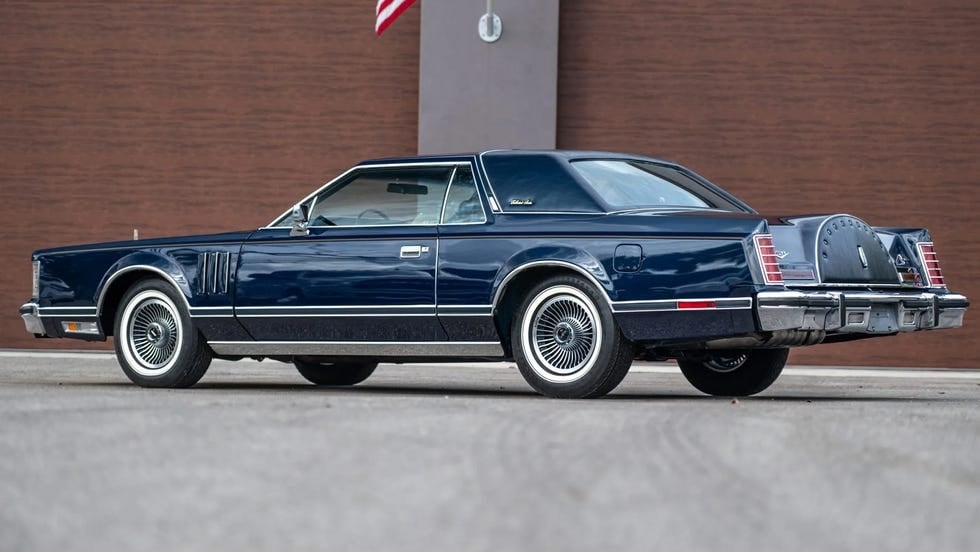 Sudut kanan belakang mobil coupe biru tua di depan dinding coklat.