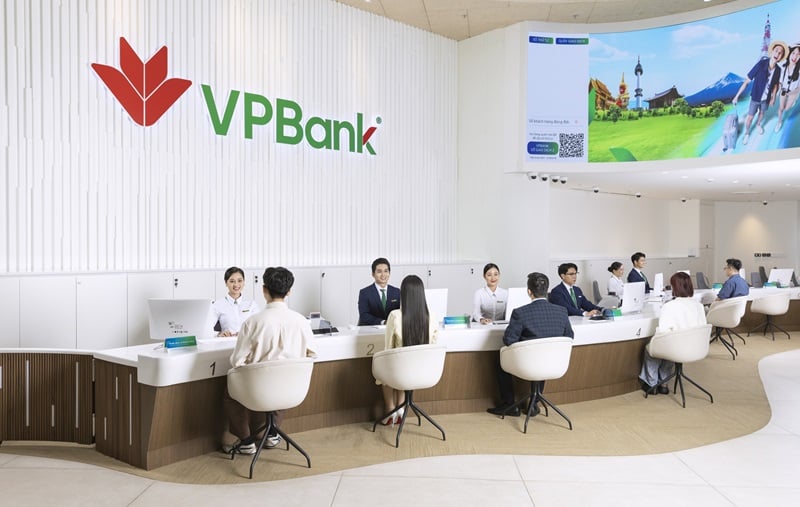 A modern and brightly lit VPBank branch interior with the large VPBank logo on a white wall. Several bank tellers are assisting customers at a long, curved service counter, while a large digital screen displays travel imagery in the background.